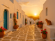 Coucher de soleil sur une ruelle typique de Sifnos, maisons blanches, portes bleues, pots de fleurs rouges et chat assis sur un muret.