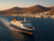 Ferry approchant du port d’une île des Cyclades au coucher du soleil, avec maisons blanches en bord de mer et collines dorées en arrière-plan.