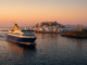 Ferry arrivant au port de Naxos au lever du soleil, avec la Portara antique et les maisons blanches éclairées par une lumière dorée.