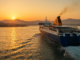 Ferry blanc et bleu quittant le port du Pirée au lever du soleil, avec Athènes et ses collines en arrière-plan, éclairé par une lumière dorée sur la mer Égée