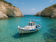 Petit bateau de pêche blanc et bleu amarré dans une crique aux eaux turquoise des Cyclades, entourée de falaises rocheuses sous un ciel ensoleillé.