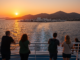 Vue depuis un ferry approchant Paros au coucher du soleil, avec des voyageurs observant l’île, ses maisons blanches et ses collines baignées de lumière dorée.