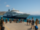 Vue réaliste d’un ferry grec à quai dans les Cyclades, sous un ciel bleu clair. Des passagers montent à bord avec leurs valises, ambiance estivale et détendue, parfaite illustration pour un article sur les trajets entre les îles.